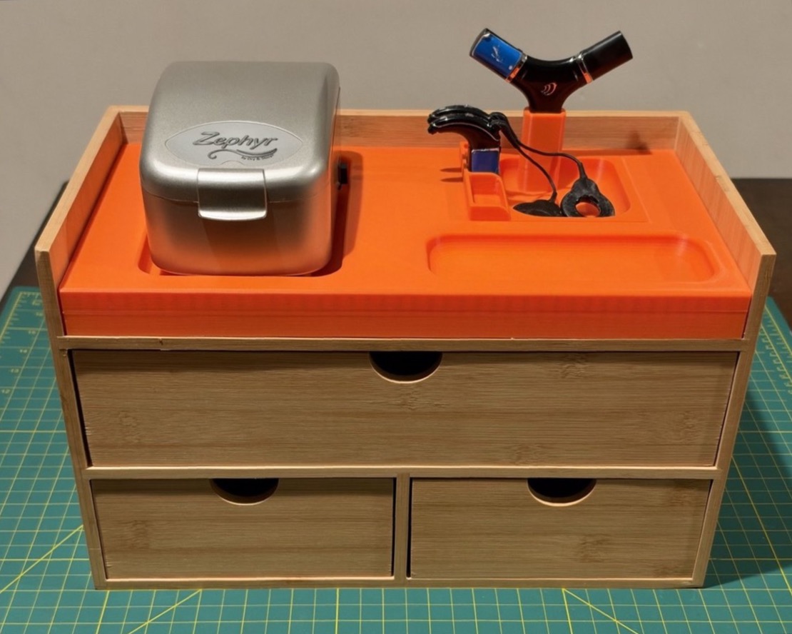 An orange 3D-printed organizer holding a cochlear implant charger, Zephyr drying station, and accessories on a wooden drawer unit.