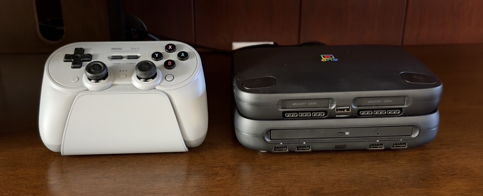 A black SuperStation One on its dock beside a white 8BitDo controller on a wooden shelf.
